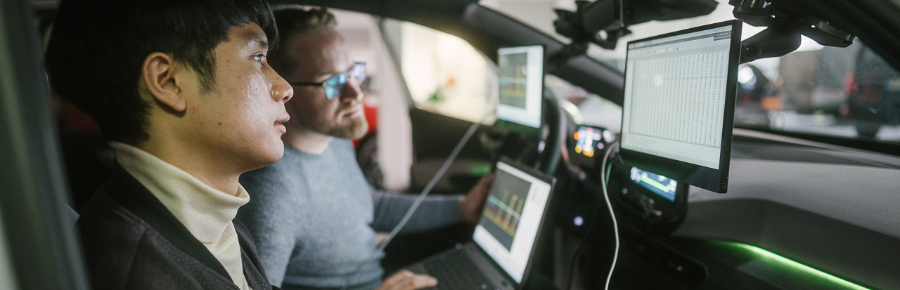 Zwei Studenten schauen in einem Pkw-Cockpit auf Monitore mit Messdaten